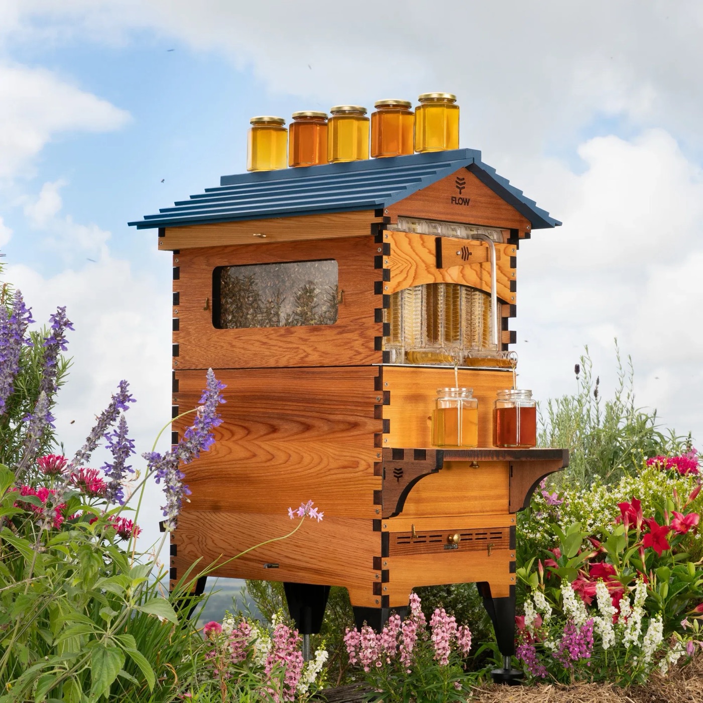 Een houten Flow Hive met een donker schuin dak, omringd door wilde bloemen en lavendel.